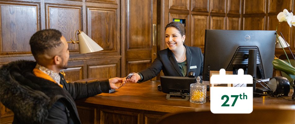 Two people sitting at a desk with a woman handing a card to a man with calendar that reads 27