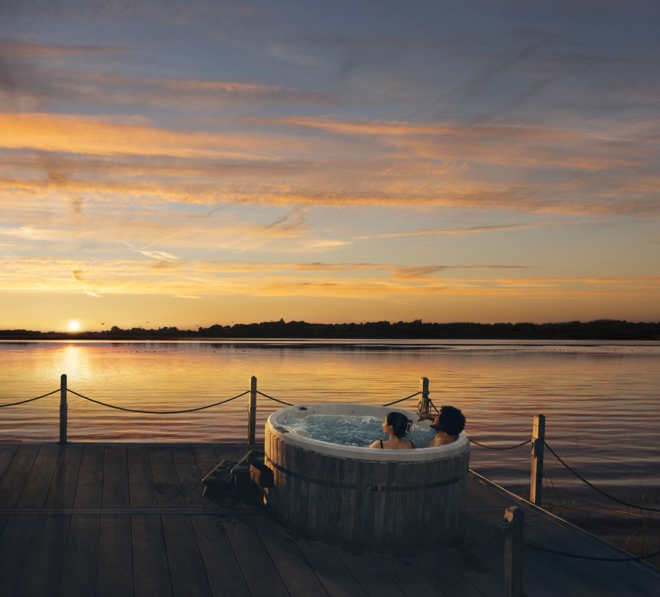 Two people sitting in a hot tub beside a lake at sunset