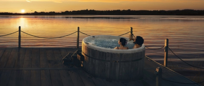 Two people sitting in a hot tub by a lake a sunset